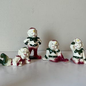 Puede incluir: Cuatro figuritas de elfos de cerámica con detalles en verde y rojo. Los elfos están en varias poses, incluyendo de pie, sentados y acostados.