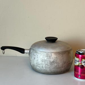 Op de afbeelding: Een zilveren aluminium pan met een zwarte handgreep en deksel. De pan heeft een versleten, getextureerd oppervlak. Een blikje Canada Dry Zero Sugar Ginger Ale staat op de achtergrond.