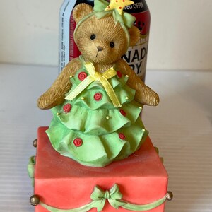 Puede incluir: Una figurita musical de &aacute;rbol de Navidad con un oso de peluche vestido con un vestido verde y amarillo. El oso lleva una estrella en la cabeza y la figurita est&aacute; sobre una caja roja con una cinta verde.