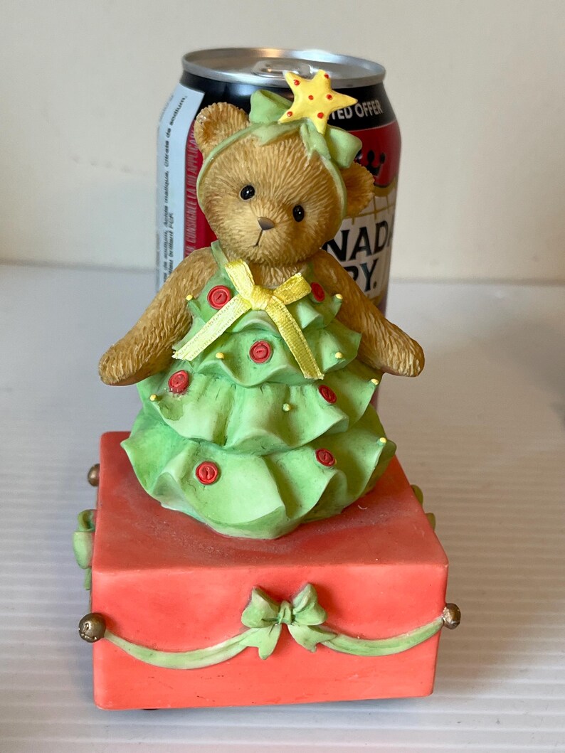 Puede incluir: Una figurita musical de &aacute;rbol de Navidad con un oso de peluche vestido con un vestido verde y amarillo. El oso lleva una estrella en la cabeza y la figurita est&aacute; sobre una caja roja con una cinta verde.