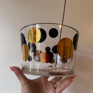 Peut inclure: Un bol en verre transparent orné d'un motif décoratif composé de cercles noirs opaques et de cercles dorés métallisés de tailles variées, répartis sur toute la surface extérieure. Le bol présente une ouverture large et une base plate.