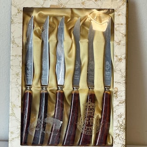 Puede incluir: Juego de seis cuchillos para carne de acero inoxidable con mangos de madera marrón en una caja blanca con detalles dorados. La caja tiene el texto "Grant Cutlery Superior Stainless Steel" impreso en ella.
