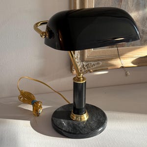 May include: A banker lamp featuring a glossy black arched shade, a gold-tone metal arm, and a cylindrical black post set upon a circular black marble base. The lamp includes a gold-tone pull chain and a yellow electrical cord with a plug.