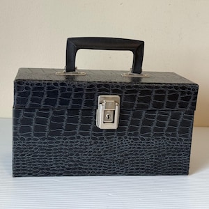 Puede incluir: Una caja rectangular negra con un patrón texturizado similar al de un cocodrilo. Cuenta con un asa negra y un cierre plateado. La caja está diseñada para el almacenamiento y tiene una estética clásica y vintage.