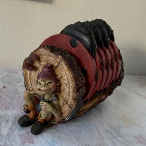 Peut inclure: Un porte-dessous de verre décoratif en forme de bûche évidée avec un extérieur en écorce brune texturée. Une petite créature verte avec un chapeau violet est assise à l'avant. Le support contient plusieurs dessous de verre rouges ronds.