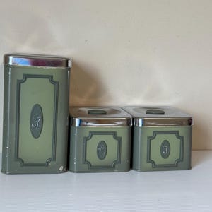May include: A trio of vintage metal canisters in shades of green with silver lids. Each canister features a decorative frame and a letter. The canisters are of different sizes, ideal for kitchen storage and measure in cm.