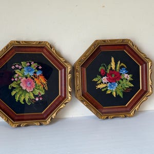 May include: Two framed floral needlepoint pictures. Each displays a bouquet of colourful flowers on a black background. The frames are octagonal with a gold-tone outer edge and a reddish-brown inner edge.