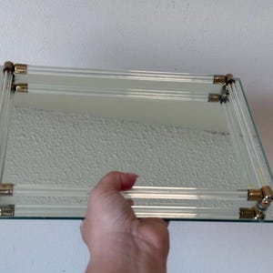 以下が含まれることがあります： 透明なアクリルチューブと金色の金属コーナーアクセントが付いた長方形のガラス棚。手で持っており、反射面とすっきりとしたラインが特徴です。デザインはヴィンテージまたはレトロな美学を示唆しています。