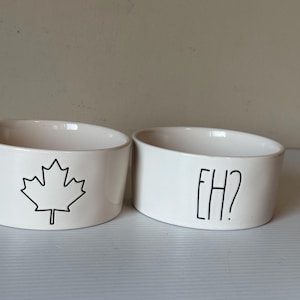 May include: Two white ceramic bowls. One bowl features a black outline of a maple leaf, and the other has the text "EH?" in black. The bowls are oval-shaped and suitable for pet food or water.