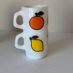 Może przedstawiać: Dwa białe ceramiczne kubki ułożone jeden na drugim. Każdy kubek ma czarny kontur owocu z czarnym liściem. Górny kubek ma pomarańczę, a dolny kubek ma żółtą cytrynę.