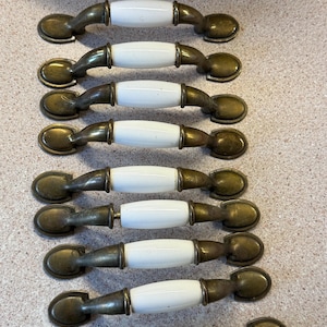 Peut inclure: Un ensemble de neuf poignées de placard, chacune dotée d'un centre en céramique blanche lisse flanqué de deux extrémités en métal couleur laiton antique. Les pièces sont disposées en colonne verticale, avec un design incurvé et des plaques ovales.