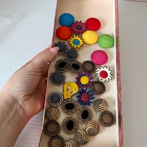 以下が含まれることがあります： さまざまな色のボタンが詰まった箱。丸いボタン、花の形をしたボタン、複雑なデザインの金属ボタンなどがあります。一部のボタンには漫画のキャラクターが描かれており、他のボタンには黒い中心があります。