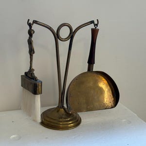 Peut inclure: Un ensemble d'outils de cheminée en laiton comprenant un support avec un détail circulaire au sommet, une brosse avec un manche métallique et des poils blancs, et une pelle avec un manche en bois foncé suspendus aux bras incurvés.