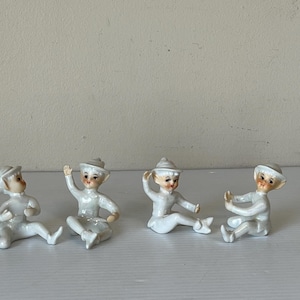 Puede incluir: Cuatro pequeñas figuras de cerámica blanca de figuras sentadas con sombreros. Cada figura tiene una cara pintada con mejillas rosadas y posa de manera diferente. Las figuras están sobre una superficie blanca.