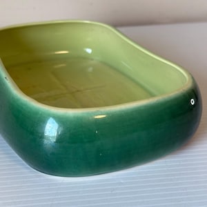May include: A ceramic, rectangular dish with rounded edges. The exterior is a glossy, deep green, while the interior is a lighter, yellowish-green. The dish has a smooth, reflective surface, suggesting it is glazed.