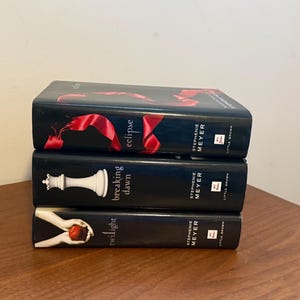 Puede incluir: Una pila de tres libros de tapa dura de Stephenie Meyer con lomos negros. El libro superior es Eclipse con una cinta roja, el del medio es Breaking Dawn con una pieza de ajedrez blanca y el inferior es Twilight con manos sosteniendo una manzana roja.