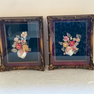 Peut inclure: Deux cadres muraux rectangulaires avec des bordures dorées ornées et des fonds en velours sombre. Chaque cadre contient un arrangement floral en trois dimensions composé de divers coquillages roses, blancs et jaunes sur une base en coquillage.