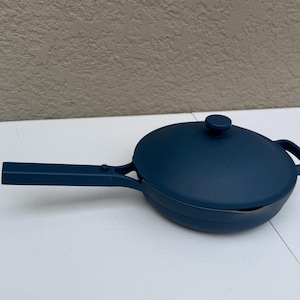 Puede incluir: Una sartén antiadherente de color azul profundo con tapa a juego. La sartén presenta un mango largo y recto con un pequeño botón circular, un cuerpo redondeado y una segunda asa pequeña en el lado opuesto. La tapa tiene un pomo central.