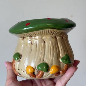 Peut inclure: Un récipient en céramique en forme de champignon, avec un dessus vert parsemé de points rouges. Le corps est beige avec des lignes verticales, et de petits champignons verts et jaunes ornent la base. Une pièce décorative fantaisiste.