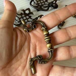 Puede incluir: Primer plano de tiradores de cajón de latón antiguo con diseños ornamentados. Los tiradores tienen un asa curvada y detalles decorativos, con un acabado cálido y envejecido. Se ven varios tiradores, lo que sugiere un juego para la restauración de muebles.