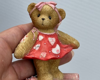 May include: A small, brown teddy bear figurine wearing a red dress with pink hearts and a pink bow. The bear is standing on a pink and white fabric base with a spool of thread and a needle.