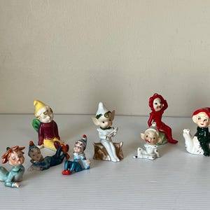 Puede incluir: Una colección de figuras de elfos de cerámica en varias poses. Los elfos visten trajes coloridos, incluyendo rojo, verde, azul y amarillo. Algunos están sentados, tumbados o sosteniendo objetos. Las figuras son probablemente artículos vintage o de colección.