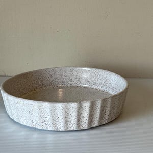 May include: A speckled, off-white ceramic pie dish with a fluted edge. The round dish has a shallow depth and a speckled pattern throughout. The dish is suitable for baking and serving pies, quiches, or other baked goods.