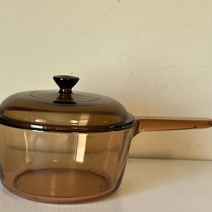 May include: A translucent amber-coloured glass saucepan with a matching glass lid. The pot features a single integrated glass handle extending from the side and a circular knob on the centre of the lid. The glass is smooth and uniform in colour.