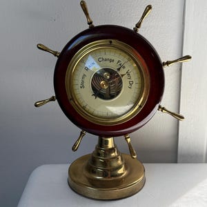 May include: A nautical-style barometer featuring a dark mahogany-colored circular frame with brass spokes resembling a ship's wheel. The central gauge displays the text Stormy, Rain, Change, Fair, and Very Dry with a brass base stand.