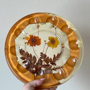 Pode incluir: Um cinzeiro redondo de resina transparente com uma borda dourada e um design de flores prensadas. As flores são amarelas e laranja.
