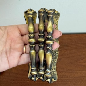 Könnte beinhalten: Drei Schubladengriffe aus messingfarbenem Metall mit einem zentralen Spindeldesign, dekorativen seilartigen Details und verzierten, strukturierten rechteckigen Rückplatten mit spitz zulaufenden Enden.