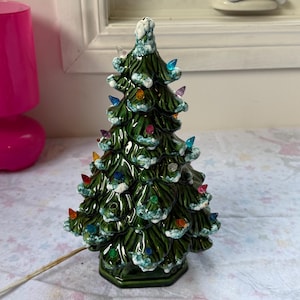Könnte beinhalten: Eine Weihnachtsbaumfigur aus Keramik in glänzendem Dunkelgrün mit weißer, schneeartiger Textur an den Zweigen. Der Baum ist mit kleinen, bunten Kunststoff-Lämpchen in den Farben Blau, Rot, Orange, Violett und Transparent verziert.