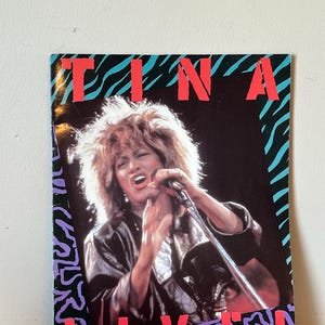 May include: A concert poster featuring a photo of Tina Turner singing into a microphone. The poster has a black background with a zebra print border and the words "Tina Live!" in red.