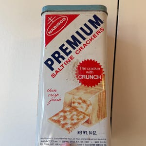 May include: A vintage rectangular metal Nabisco Premium Saltine Crackers tin with a white background, red and blue text, and a graphic of crackers. The tin features the text The cracker with crunch, thin crisp fresh, and a net weight of 14 oz.