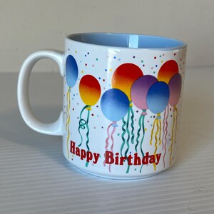 Può includere: Tazza in ceramica bianca con bordo blu e un design di palloncini colorati. La tazza ha la scritta "Happy Birthday" stampata sul davanti.