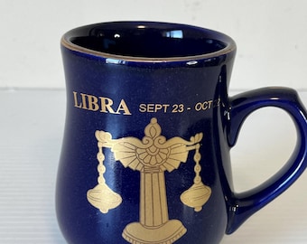 Pode incluir: Uma caneca de cerâmica azul escura com um símbolo de Libra e texto dourados. A caneca tem uma alça e as palavras "LIBRA SEPT 23 - OCT 22" em letras douradas. O design inclui um símbolo de balança.