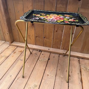 May include: A vintage black serving tray with a floral design, resting on a gold-colored folding stand. The tray features a scalloped edge and a colorful arrangement of pink, yellow, and orange flowers. The stand is made of thin, curved metal.