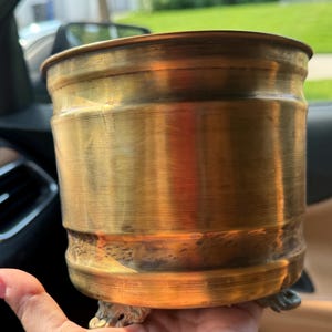 Puede incluir: Una maceta cilíndrica de latón con un acabado metálico pulido y detalles acanalados horizontales a lo largo del cuerpo. El recipiente presenta un borde superior ancho y abierto y se apoya sobre tres pies pequeños, ornamentados y claros.