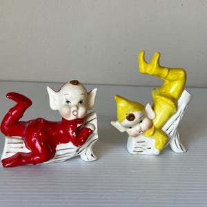 Puede incluir: Un par de figuras de elfos de cerámica vintage. Uno es rojo, el otro amarillo. Ambos están acostados sobre troncos blancos y dorados. Los elfos tienen piel blanca, orejas puntiagudas y sonríen. Un artículo decorativo caprichoso.
