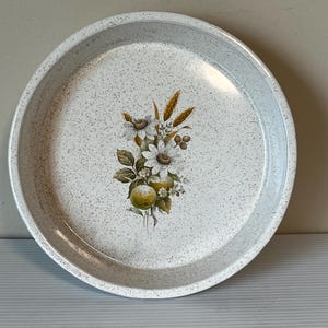 May include: A round serving tray in white with a speckled design. It features a floral pattern with white daisies, wheat, and green apples. The tray has a raised edge and is suitable for serving food or displaying items.