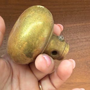 May include: A round, metallic brass door knob with a smooth, weathered surface. The knob features a cylindrical neck with a small, visible metal screw and a circular opening on the side, designed for attachment to a door mechanism or spindle.