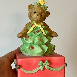 Puede incluir: Una figurita musical de &aacute;rbol de Navidad con un oso de peluche vestido como un &aacute;rbol de Navidad. El oso lleva un vestido verde con una cinta amarilla y una estrella amarilla en la cabeza. La figurita est&aacute; sobre una base roja con una cinta verde y un lazo.