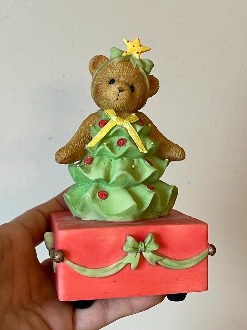 Puede incluir: Una figurita musical de &aacute;rbol de Navidad con un oso de peluche vestido como un &aacute;rbol de Navidad. El oso lleva un vestido verde con una cinta amarilla y una estrella amarilla en la cabeza. La figurita est&aacute; sobre una base roja con una cinta verde y un lazo.