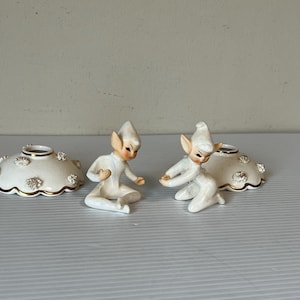 Puede incluir: Dos figuras de elfos de cerámica con sombreros puntiagudos y mejillas rosadas, arrodilladas cerca de dos candelabros a juego. Los candelabros son blancos con adornos dorados y detalles florales. Las figuras están en una pose juguetona.