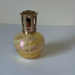 May include: A decorative, pear-shaped oil lamp with a gold-coloured metal top. The glass body has a yellow and cream swirl pattern with iridescent highlights. The lamp is designed for home fragrance.