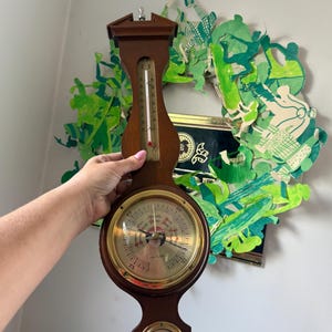 May include: A wall-mounted weather station featuring a dark wood frame with a vertical thermometer, a large central brass-rimmed barometer with a dial labeled Solar, and a smaller brass-rimmed hygrometer at the bottom with a needle indicator.