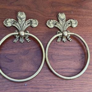 Peut inclure: Une paire d'anneaux porte-serviettes en laiton avec une fixation murale décorative en forme de fleur de lys sur le dessus. Chaque anneau circulaire est fixé à une plaque de base en métal en forme de lys stylisé avec des détails complexes.