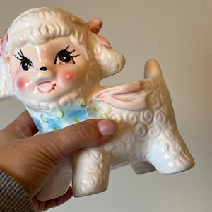 Op de afbeelding: Een witte keramische lammenpot met roze en blauwe bloemdetails. Het lam heeft een roze strik en een geschilderd gezicht met gesloten ogen en een roze tong die uitsteekt.