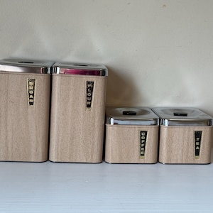 May include: A collection of four vintage kitchen canisters with a light wood grain effect and silver lids. The canisters are labelled "SUGAR", "FLOUR", "COFFEE", and "TEA" in gold lettering. The set is arranged on a white surface.