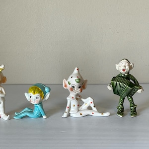 Puede incluir: Cuatro figuras de elfos de cerámica en varias poses. Uno viste un traje blanco con un sombrero amarillo, otro está con un traje azul, un tercero con un traje blanco con lunares rojos, y el último toca el acordeón.